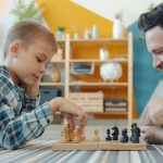 Smart child playing chess with dad at home lying on floor together having fun with intellectual sports game. Happy family and leisure activity concept.
